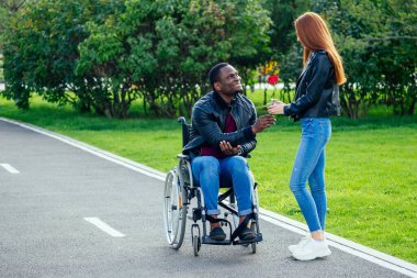 Tekerlekli sandalyede oturan Afro-Amerikan bir adam, kızıl saçlı kız arkadaşı sonbahar parkında bebek arabası sürüyor. Romantik bir randevu.