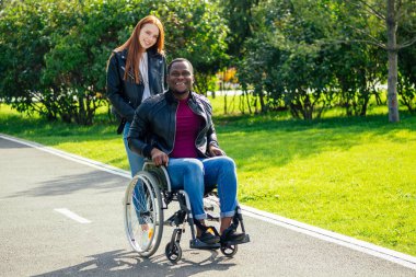 Tekerlekli sandalyede oturan Afro-Amerikan bir adam, kızıl saçlı kız arkadaşı sonbahar parkında bebek arabası sürüyor. Romantik bir randevu.