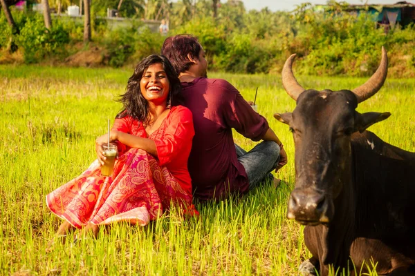 Счастливая индийская пара влюбленная сидит на поле возле коровы, пьет тростниковый сок на ферме Гоа — стоковое фото