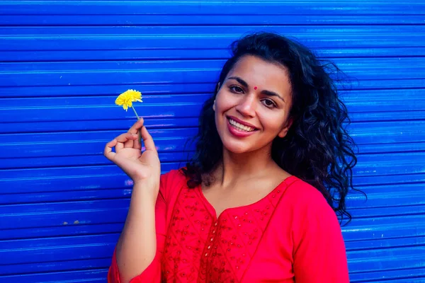 Atractiva hermosa feliz joven latina hispana con bindi de teca en la frente sonriendo en fondo azul de la calle de la pared sosteniendo flover fresco —  Fotos de Stock