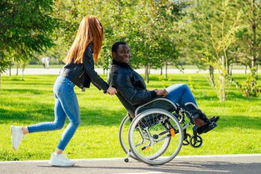 Tekerlekli sandalyede oturan Afro-Amerikan bir adam, kızıl saçlı kız arkadaşı sonbahar parkında bebek arabası sürüyor. Romantik bir randevu.