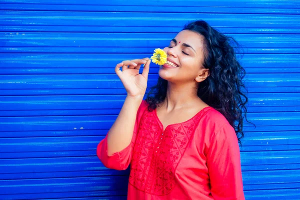 Atractiva hermosa feliz joven latina hispana con bindi de teca en la frente sonriendo en fondo azul de la calle de la pared sosteniendo flover fresco —  Fotos de Stock