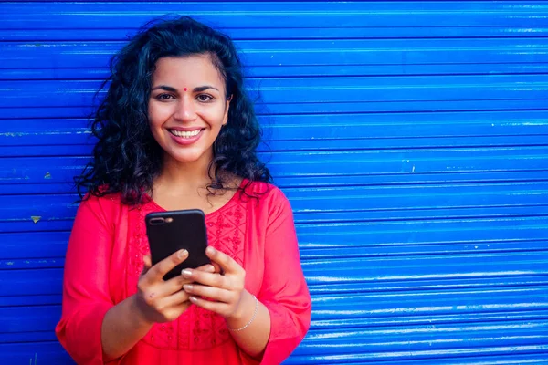 Atractiva hermosa feliz joven latina hispana mujer con bindi de teca en la frente sonriendo en la pared azul calle fondo celebración de teléfono inteligente moderno —  Fotos de Stock