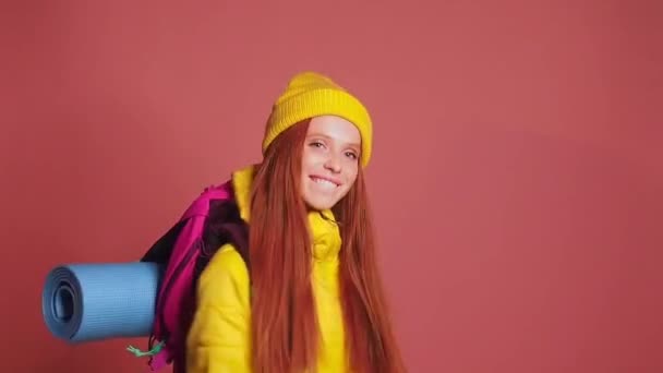 ピンクのスタジオの背景にある赤い髪の生姜の女性は、帽子と黄色のウインドブレーカージャケットを着て、バックパックを肩に持っています。彼女は行く準備ができている — ストック動画