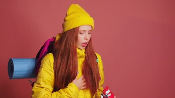 Tired redhaired ginger woman in pink studio background wearing yellow windbreaker jacket with hat and holding on her back shoulders the backpack ,she feeling thirst — Stock Video
