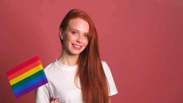 Redhaired mulher gengibre em rosa estúdio fundo acenando arco-íris bandeira colorida — Vídeo de Stock