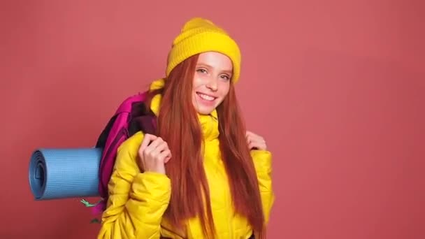 Pelirroja de jengibre en el fondo del estudio de color rosa con chaqueta cortavientos amarillo con sombrero y sosteniendo sobre sus hombros la mochila. ella está lista para irse — Vídeos de Stock