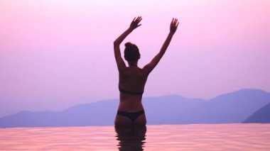 Genç siluet kadın sonsuz yüzme havuzunda oturuyor ve iki elini sallıyor. Günbatımının tadını çıkarıyor.