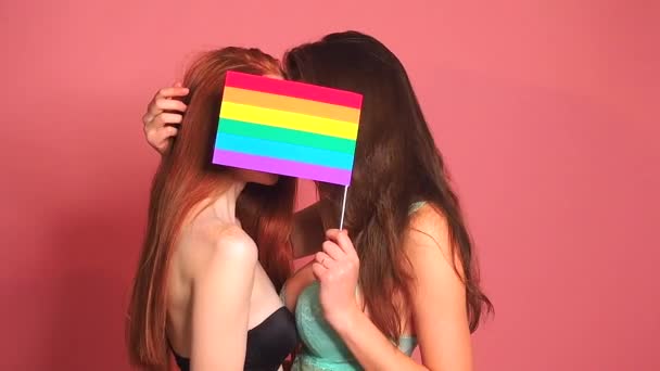 Pelirroja pelirroja mujer en rosa fondo del estudio besándose y escondiéndose detrás de la bandera del arco iris — Vídeo de stock