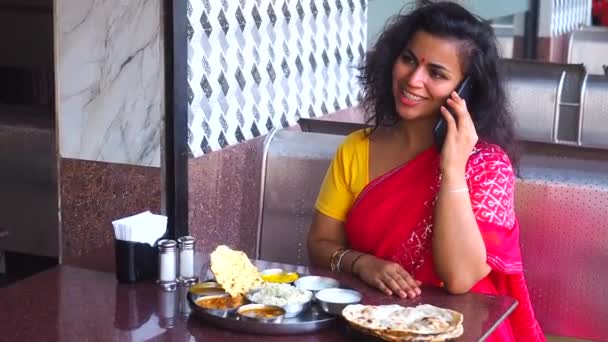 Hermosa mujer comiendo dosa chutney naan y tali indio y hablando por teléfono en un restaurante.She vistiendo sari rojo con pendientes de oro, con tilaka en la frente — Vídeo de stock