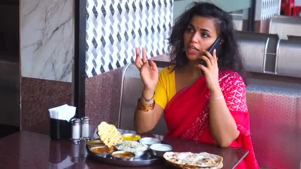 Gyönyörű nő eszik dosa chutney naan és indiai tali és telefonon beszél egy étteremben.Ő visel piros szári arany fülbevaló, tilaka a homlokán — Stock videók