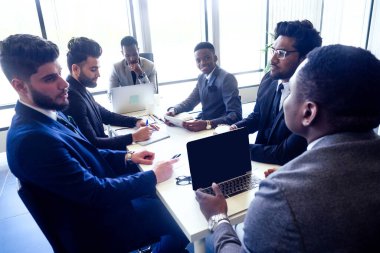 Arap ve Hindistanlı ortaklarla konuşan afro iş adamları, çok ırklı bir ekip, bir dizüstü bilgisayarla masada oturuyor ve ofiste panorama pencereli belgeler var.