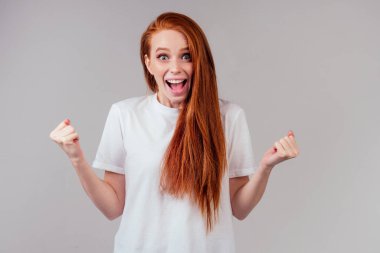 Kızıl saçlı, beyaz pamuklu kadın parmaklarını çapraz yapıp stüdyonun gri arka planında en iyisini diliyor.