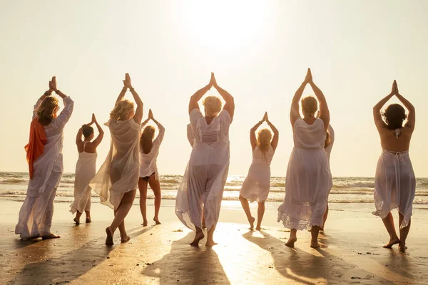 Группа молодых женщин в белом платье практикующих йогу на берегу моря — стоковое фото