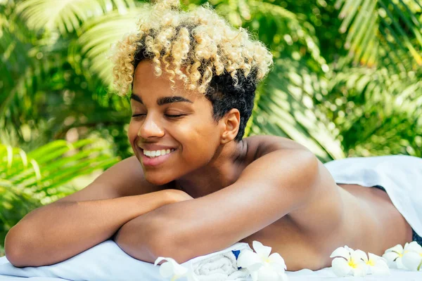 Piękna brazylijska uśmiechnięta dziewczyna leżąca na stole z ręcznikiem i białymi kwiatami, masująca się i rozpieszczająca w luksusowym centrum spa na Bali — Zdjęcie stockowe