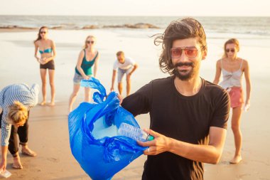 Önde oturan yakışıklı Hintli adam kum torbasından plastik topluyor. Karışık ırk arkadaşları Goa Hindistan plajında atık topluyor.