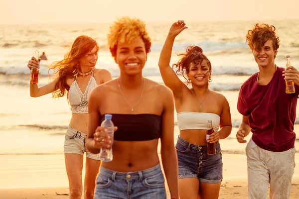 Afrikanisch-amerikanische Frau mit Plastikflasche Wasser, hinter ihrer Gruppe gemischter internationaler Freunde, die am Strand bei Sonnenuntergang mit Limonade tanzen und Spaß haben — Stockfoto