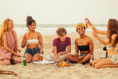 Karışık ırk çok uluslu arkadaş grubu sosis pişiriyor ve gün batımında kumda limonata içiyor.