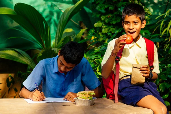 Chico de la escuela india escribiendo en el bloc de notas haciendo la tarea, buscando concentración. Su amigo con mochila se sienta en la mesa y se come una manzana en el parque — Foto de Stock