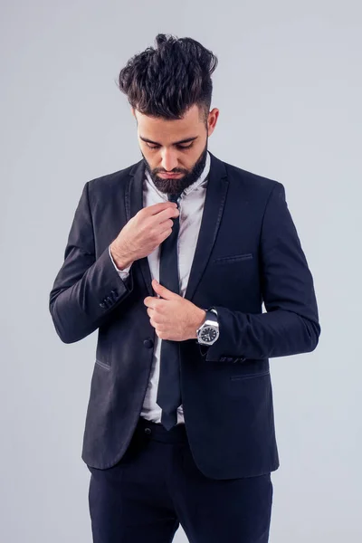 Joven turco guapo negro cabello hombre en elegante traje de negocios anudando la corbata en estudio aislar fondo blanco — Foto de Stock