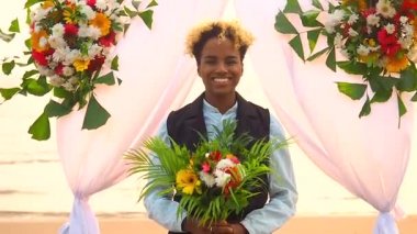 Siyah takım elbiseli Afro-Amerikan kadın damat ve düğün çiçekli kemerin altındaki tropikal plajda resmi giyinmiş beyaz gelin.
