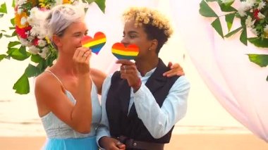 Siyah takım elbiseli Afro-Amerikan kadın damat ve tören elbisesi giymiş beyaz gelin düğün çiçekleri kemerinin altındaki tropik plajda ellerinde gökkuşağı kalbini tutuyorlar.