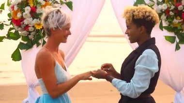Siyah takım elbiseli Afro-Amerikan kadın damat ve tören elbisesi giymiş beyaz gelin düğün kemerinin altında tropikal plajda yüzüklerini değiş tokuş ediyorlar.
