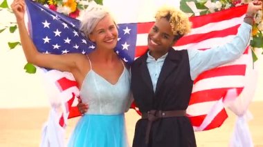 Siyah takım elbiseli Afro-Amerikan kadın damat ve mutlu Kafkasyalı sarışın gelin Amerika bayrağına sarılmış çiçek kemerinin altında