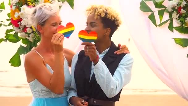 Afroamericano sposo donna in abito nero e sposa caucasica in abito da cerimonia tenendo cuore arcobaleno in mano sulla spiaggia tropicale sotto arco flover matrimonio — Video Stock