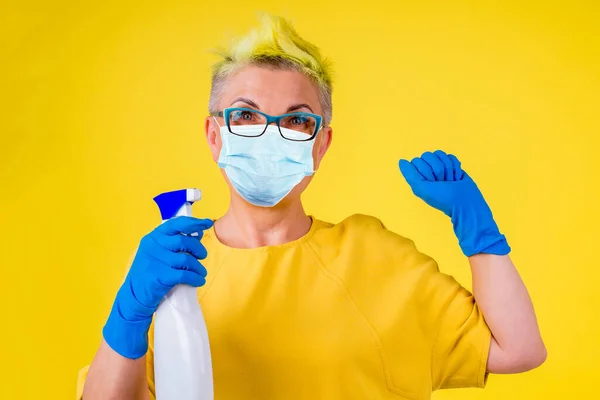 Olgun bir kadın. Renkli, sarı saçlı, kalın elbiseli ve sağlık maskeli. Arka planda yerleri yıkıyor. — Stok fotoğraf