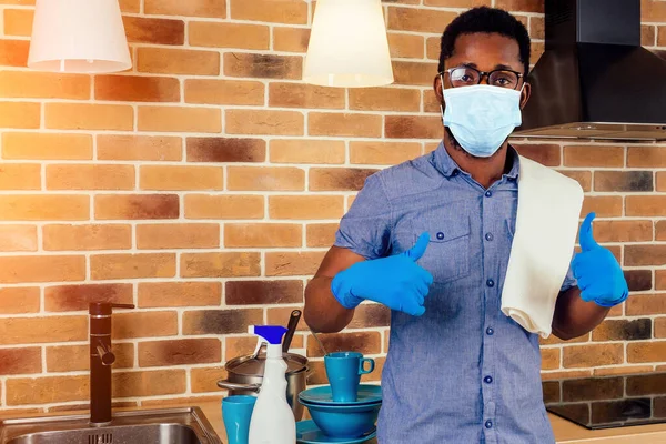 Αφρικανός άνδρας σε ιατρική μάσκα καθαρισμού εστία εστία μαγειρέματος στο σπίτι, τούβλο τοίχο φόντο — Φωτογραφία Αρχείου