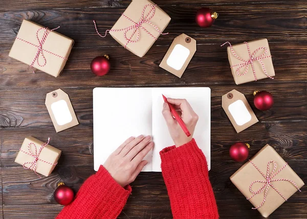 Karácsonyi ajándék listát írásban a papír, a fa háttér, ajándékok és a címkék női kezek — Stock Fotó