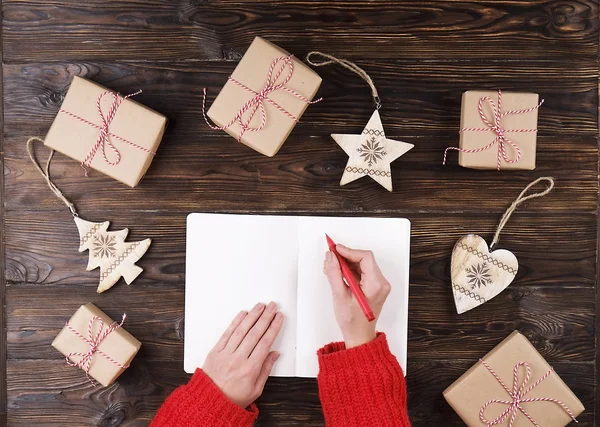 Karácsonyi ajándék listát írásban a papír, a fa háttér, ajándékok és a címkék női kezek — Stock Fotó