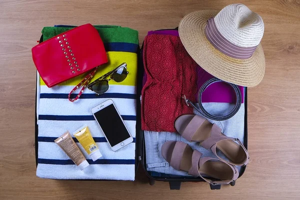 Open packed suitcase with female summer clothes and accessories, hat, sunglasses, beach towel, sunscreen and bathing suit on wooden background top view, close up — Stock Photo, Image