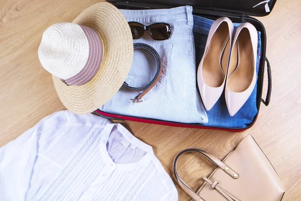 Maleta abierta con sombrero de mujer casual, gafas de sol, camisa blanca, zapatos, bolso en vista superior de piso de madera de cerca. Embalaje bolsa de viaje para concepto de viaje —  Fotos de Stock