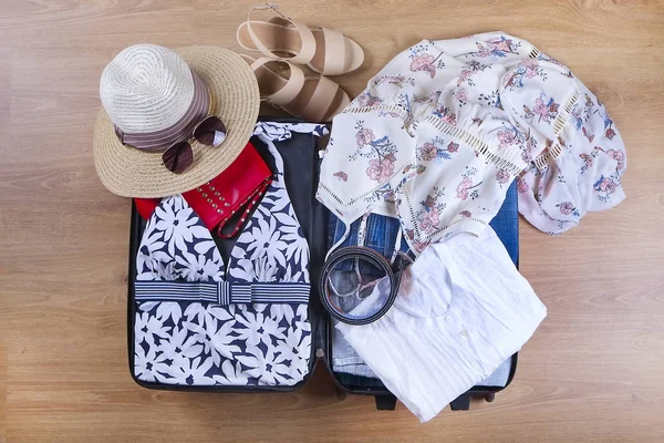 Open packed suitcase with female summer clothes and accessories, bathing suit, hat, sunglasses, sneakers, white shirt on wooden floor top view — Stock Photo, Image