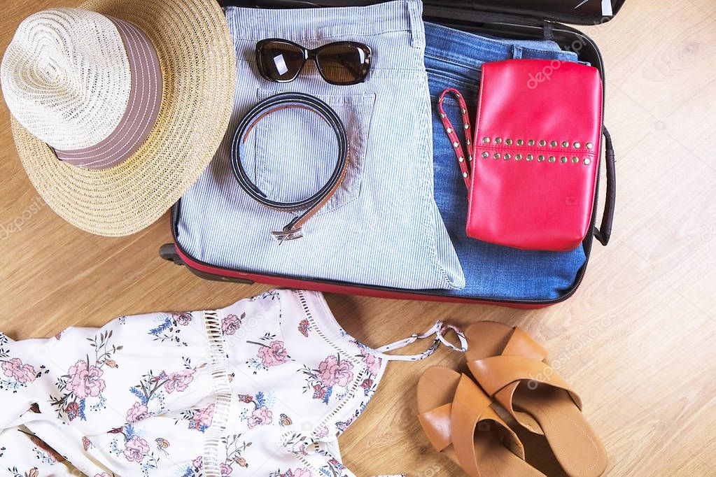 Open suitcase with casual female clothes hat, sunglasses, dress, shoes, on wooden floor top view close up. Packing travel bag for trip concept