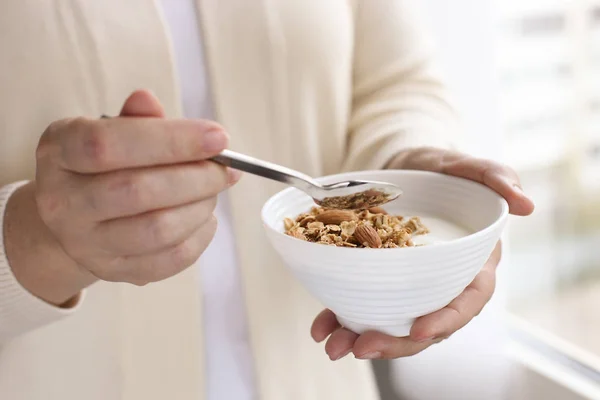 Woman eating homemade granola breakfast cereals with mixed nuts, oats and Greek yogurt in bowl. Female hands holding healthy vegetarian superfood with cashew, almond, hazelnut. Morning light, close up — Stock Photo, Image