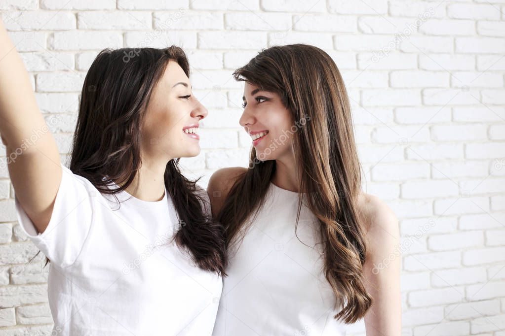 Portrait of two millennial females, fooling around in front of mobile smartphone camera. Brown eyed model girls with long brunette wavy hair and beautiful smiles in white blank shirt.