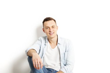 Yakışıklı genç model oturma, beyaz duvar arka planda, geniş bir gülümseme poz. Hipster rahat sokak tarzı, düz açılarak mavi gömlek.