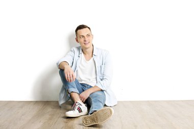 Yakışıklı genç model oturma, beyaz duvar arka planda, geniş bir gülümseme poz. Hipster rahat sokak tarzı, düz açılarak mavi gömlek.