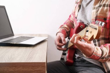 Raslantı sakal genç adam gitar şarkıları evde odada ile giyinmiş. Masa üstünde laptop. Online gitar dersleri kavramı. Erkek gitarist pratik akor kulpları.