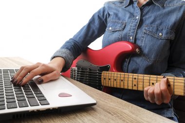 Raslantı giyimli genç kadın şarkıları evde odada gitar ile. Masa üstünde laptop. Online gitar dersleri kavramı. Erkek gitarist pratik akor kulpları.