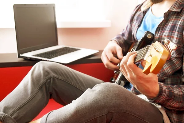 Terloops gekleed jonge man met gitaar spelen van liederen in de ruimte thuis. Online gitaar lessen concept. Mannelijke gitarist beoefenen snaar grepen. — Stockfoto