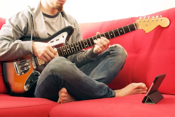 Casually dressed young man with guitar playing songs in the room at home. Online guitar lessons concept. Male guitarist practicing chord grips.