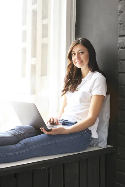 Happy millennial girl w/ laptop on windowsill. Portrait of young woman with diastema gap between teeth. Beautiful smile. Minimal interior, black brick textured wall background, loft style. Copy space.