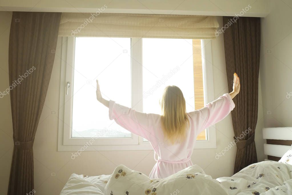 Big spacey hotel room full of sunlight and sun beams. Optimistic start of the day. Blond woman cozy home clothing welcoming the morning daylight. New day new me concept.