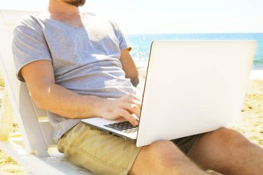 Dizüstü bilgisayarda çalışan genç adam sahilde oturan gri tişörtlü. Blogger, yazar, coder, uzak alt tarama defter üzerinde araştırma sörf. Kablosuz internet deniz kıyısında. Arka plan, kopya alanı