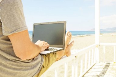 Dizüstü bilgisayarda çalışan genç adam sahilde oturan gri tişörtlü. Blogger, yazar, coder, uzak alt tarama defter üzerinde araştırma sörf. Kablosuz internet deniz kıyısında. Arka plan, kopya alanı