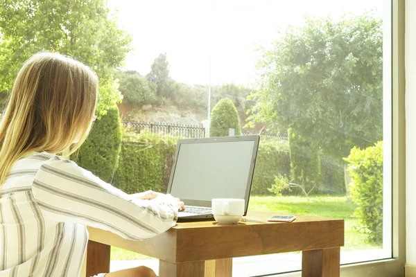 Vacker hipster kvinna i lös rand skjorta sitter hemma, med hjälp av dator. Kvinnliga författare skrivit manus hemma, av stora fönster, dricka kaffe. Kopiera utrymme, bakgrund, närbild — Stockfoto